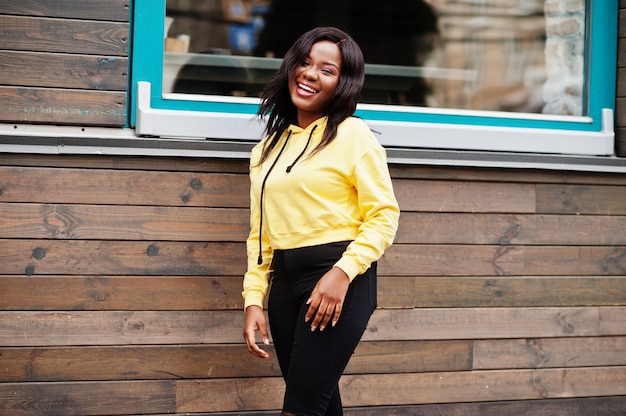 Hipster african american girl wearing yellow hoodie posing at street against wooden house with windows.