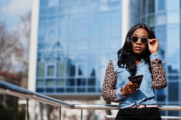Hipster afro-amerikaans meisje met een zonnebril, jeans-shirt met luipaardmouwen, houdt een mobiele telefoon op straat tegen een modern kantoorgebouw