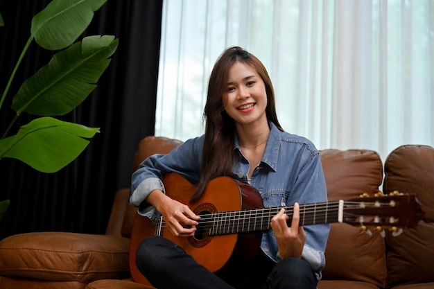 Hipster Asian female in jean jacket playing guitar un her modern vintage living room