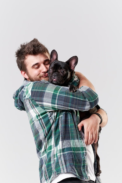 Foto ragazzo barbuto hipster che tiene e abbraccia un simpatico cane bulldog francese tra le braccia con amore e gioca con lui, contro un muro bianco