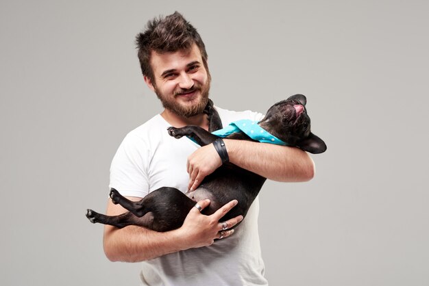 Photo hipster bearded guy holding and hugging a nice french bulldog dog in his arms with love and playing with him