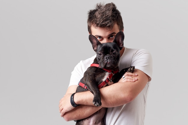 Hipster Bearded guy holding and hugging a nice French Bulldog dog in his arms with love and playing with him