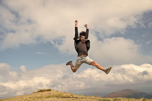 Hipster or brutal macho conquers mountain Man with brutal appearance jumps Freedom concept Man with beard enjoy freedom jumpimg on top of mountain Hipster feels free while hiking sky background