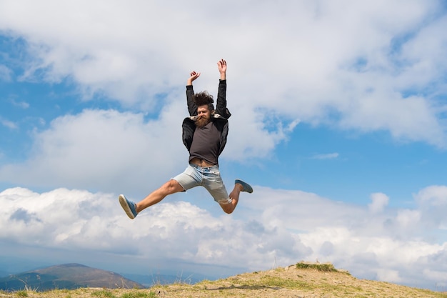 Hipster or brutal macho conquers mountain. Man with brutal appearance jumps. Freedom concept. Man with beard enjoy freedom, jumpimg on top of mountain. Hipster feels free while hiking, sky background.