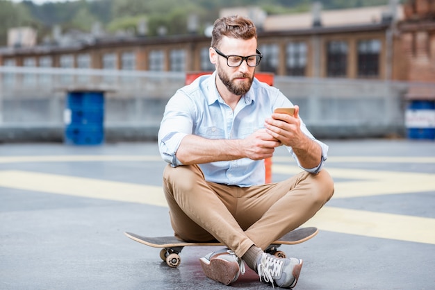 Uomo d'affari hipster seduto con uno smartphone nel parco giochi sul tetto dell'edificio industriale. concetto di stile di vita aziendale