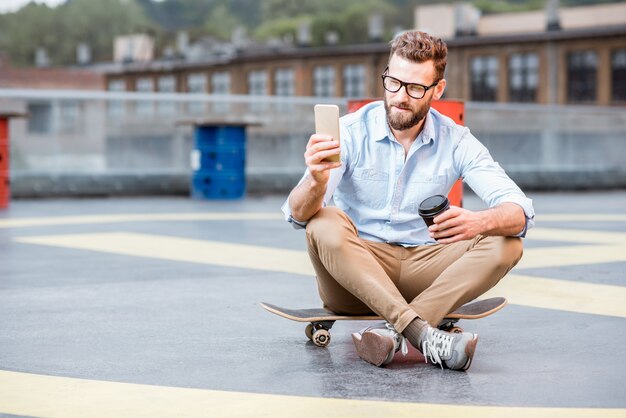 산업 건물 옥상 운동장에 스마트 폰을 들고 앉아 있는 힙스터 사업가. 라이프 스타일 비즈니스 개념