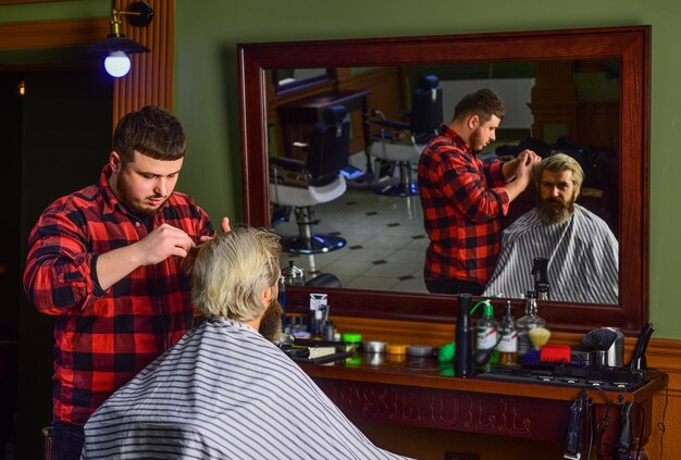 Hipster client getting haircut Barber and client Barber works hairstyle for bearded man barbershop background Guy with long dyed blond hair Professional hairstylist in barbershop interior