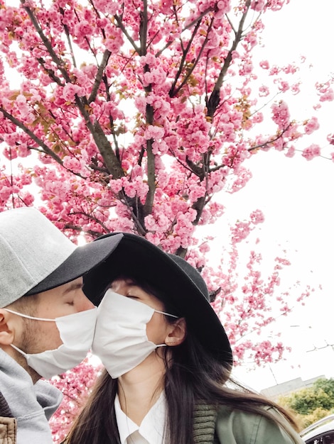 Hipster couple in face masks taking selfie and kissing on background of blooming sakura trees in city street Young family wearing medical masks during virus outbreak