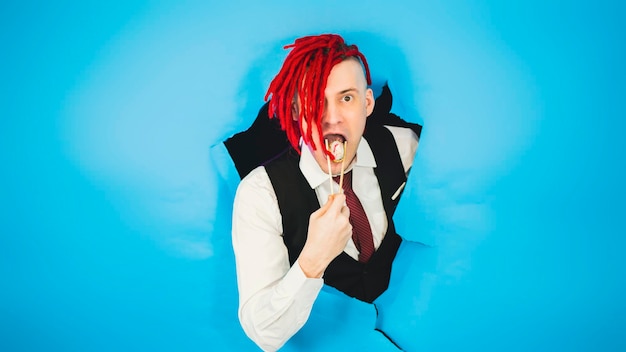 Hipster eats sushi roll man with red dreadlocks holding in hand\
sticks chopsticks with sushi roll traditional japanese food looking\
at camera through hole in blue wall in studio