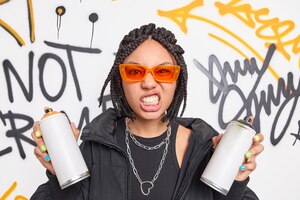 Hipster girl being street artist paints graffiti or live murales on generic wall holds spray bottles clenches teeth wears orange sunglasses black jacket and chains subculture and youth concept