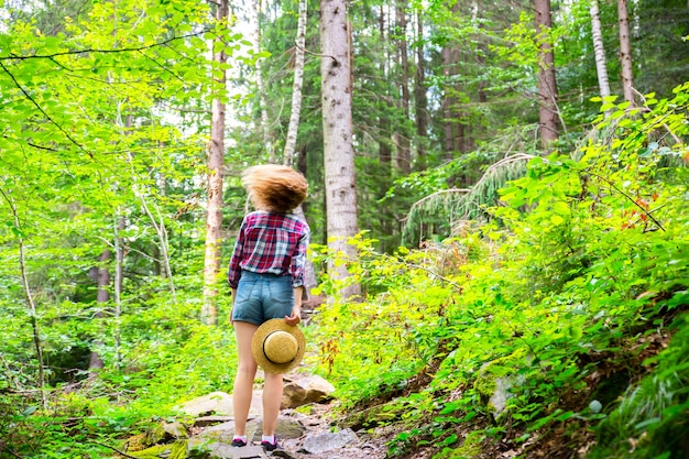 Photo hipster girl in the mountains. stylish woman in straw hat and checkered shirt in forest. wanderlust concept. freedom and psychology concept. beautiful nature.