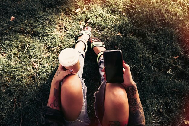 スマートフォンとコーヒーと一緒に公園に座っている流行に敏感な女の子