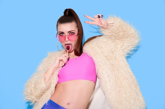 Hipster girl wearing bright colored clothes and fur coat lick lollipop and show victory gesture on blue wall