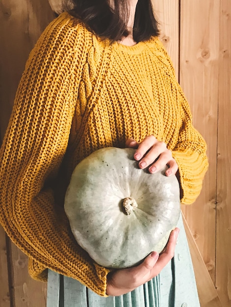 Hipster girl in yellow sweater holding pumpkins on rustic wooden background Fall rural decor and arrangement Autumn harvest Thanksgiving concept