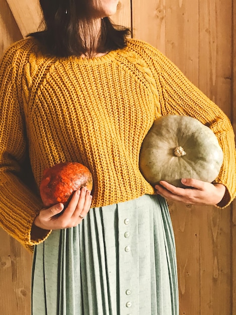 Hipster girl in yellow sweater holding pumpkins on rustic wooden background Fall rural decor and arrangement Autumn harvest Thanksgiving concept