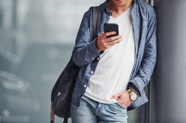 Ragazzo hipster in bei vestiti sta al chiuso con il telefono in mano