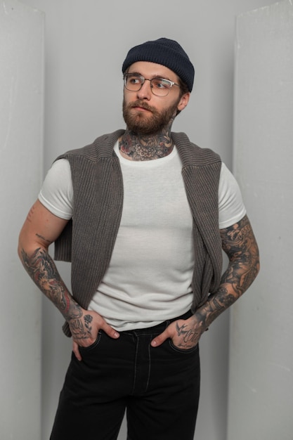 Фото Хипстер красивый крутой человек с прической и бородой с татуировкой с модными очками в стильной белой футболке и свет стоит на белом фоне в студии