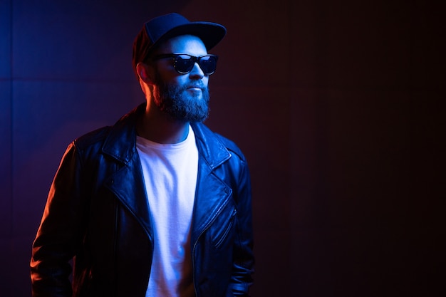 Hipster handsome man on the city streets being illuminated by neon signs. He is wearing leather biker jacket or asymmetric zip jacket with black cap, jeans and sunglasses.