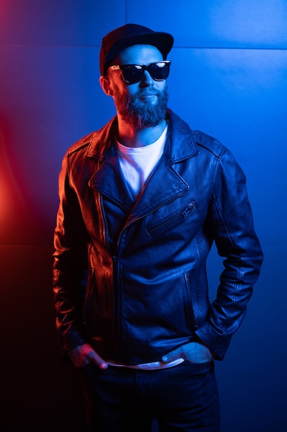 Hipster handsome man on the city streets being illuminated by neon signs. He is wearing leather biker jacket or asymmetric zip jacket with black cap, jeans and sunglasses.