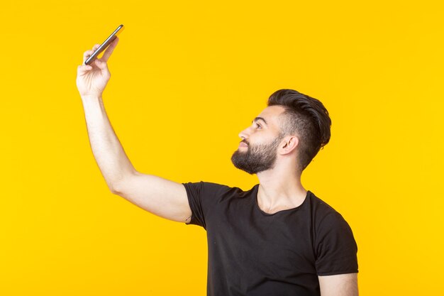 Hipster handsome young bearded man taking a selfie on his smartphone over yellow wall. concept