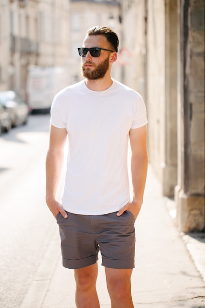 Hipster knap mannelijk model met baard, gekleed in een wit leeg t-shirt en een baseballpet met ruimte voor uw logo of ontwerp in een ongedwongen stedelijke stijl