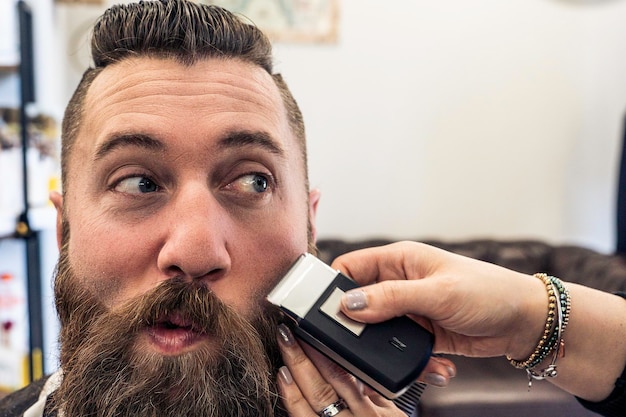 Hipster man at barbershop getting beard and hair cut Female hairdresser woman hands using electric razor to trim to classic gentleman cut Barber shop concept