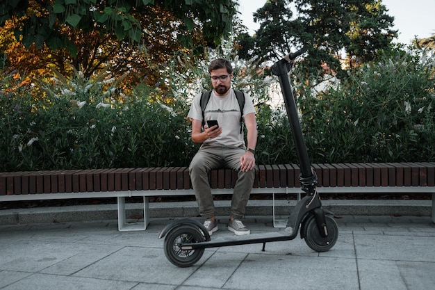 Hipster man commuter with electric scooter outdoors in city\
using smartphone