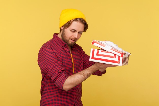 Hipster man in beanie muts en geruit hemd opening geschenkdoos gluren binnen met interesse in afwachting van leuk cadeau uitpakken verjaardag verrassing studio opname geïsoleerd op gele achtergrond