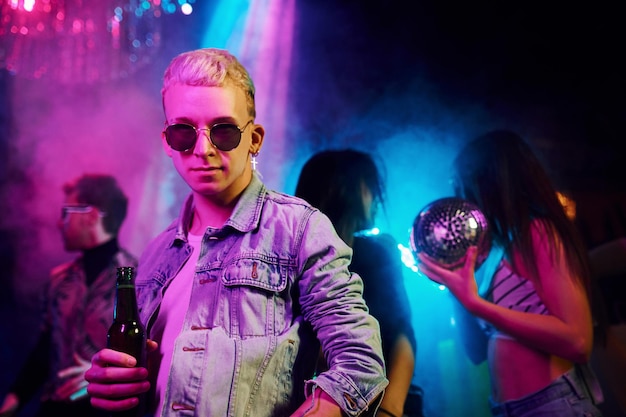 Foto hipster man in zonnebril en met fles alcohol poseren voor de camera voor jonge mensen die plezier hebben in de nachtclub met kleurrijke laserlichten