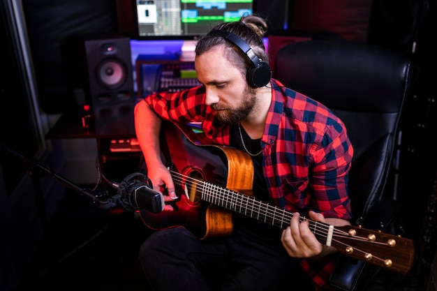 Hipster man met baard in hoofdtelefoon speelt gitaar en zingt zijn nieuwe nummer in stereostudio om een gloednieuw nummer op te nemen.