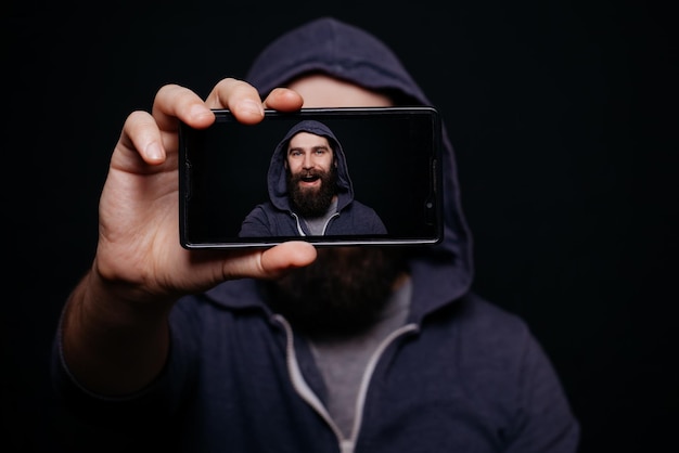 Hipster man with a beard taking picture smartphone selfportrait screen view snapshot studio on a black background