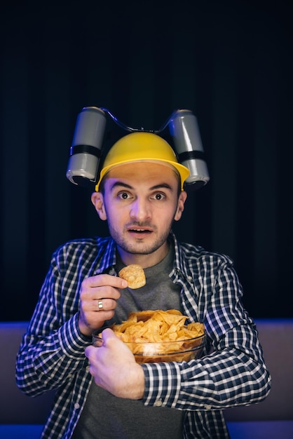 Hipster man with beer helmet on the head eating chips while watching TV At home