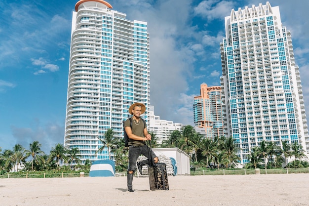 Hipster man with luggage bag on wheels standing on the south beach in miami