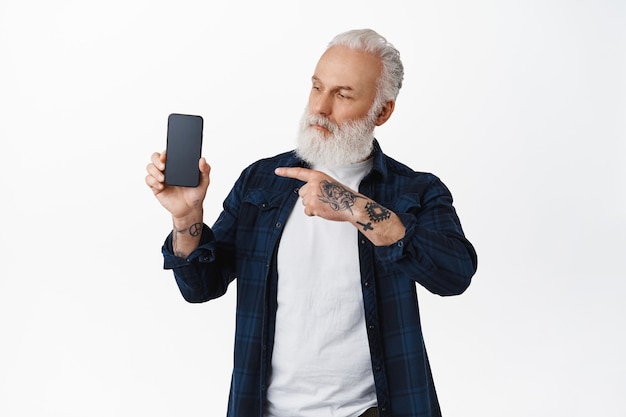 Hipster old man shows mobile phone app, pointing at smartphone screen and looking serious, recommending application, standing over white wall