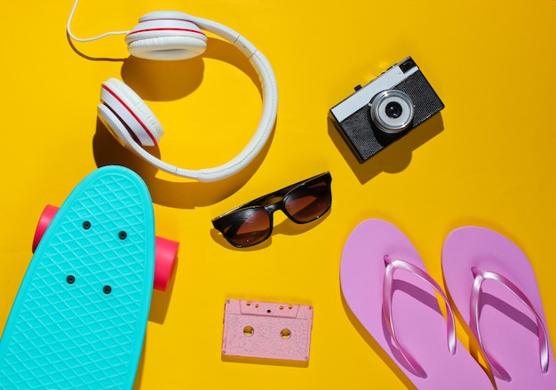 Hipster outfit. Skateboard, audio cassette, headphones, flip flop, retro camera, sunglasses on yellow background.