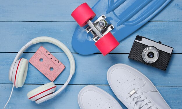 Photo hipster outfit. skateboard, headphones, audio cassette, sneakers, retro camera on blue wooden background. creative fashion minimalism. minimal summer fun. pop art. 80s. copy space. top view