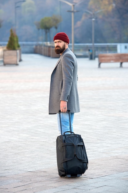 Hipster ready enjoy travel Carry travel bag Man bearded hipster travel with luggage bag on wheels Adjust living in new city Traveler with suitcase arrive airport railway station urban background
