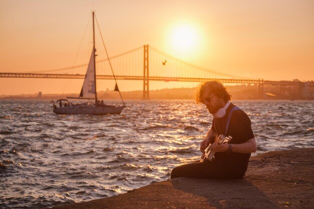 Hipster straatmuzikant in het zwart elektrische gitaar spelen in de straat bij zonsondergang aan de kade met 25 april brug en jachtboot op de achtergrond Lissabon Portugal