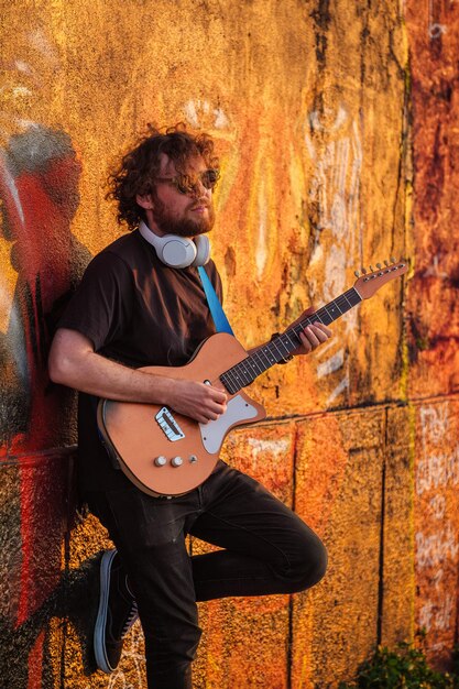 Hipster street musician in black playing electric guitar in the street on sunset leaning on a wall