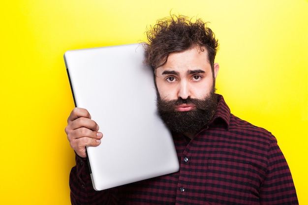 Hipster student with his laptop near the face on yellow background