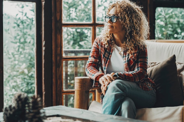 Hipster style pretty lady sitting at home on the sofa and looking outside thw windows a green forest woods. Cozy cabiness chalet female people relax lifestyle. Young adult woman enjoy leisure at home