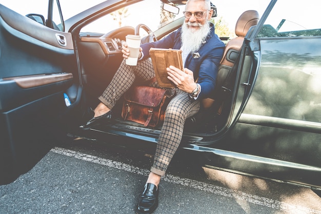 Uomo alla moda hipster utilizzando la tavoletta seduto all'interno di un'auto elettrica convertibile - imprenditore senior divertendosi con le tendenze della tecnologia - tecnologia, sistemi ecologici e concetto di moda - focus on face