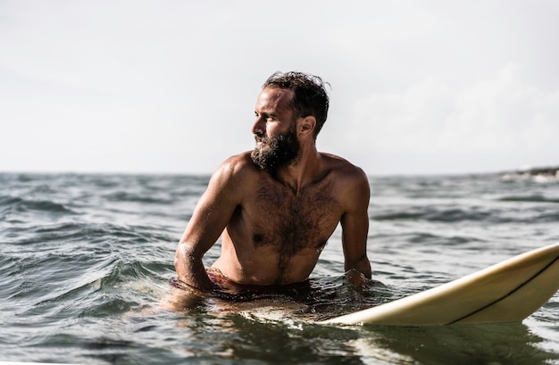 Surfista hipster seduto sulla sua tavola da surf nell'acqua dell'oceano e in attesa di una grande onda fit uomo barbuto che si allena con la tavola da surf al mare concetto di avventura e libertà facendo sport estremi acquatici