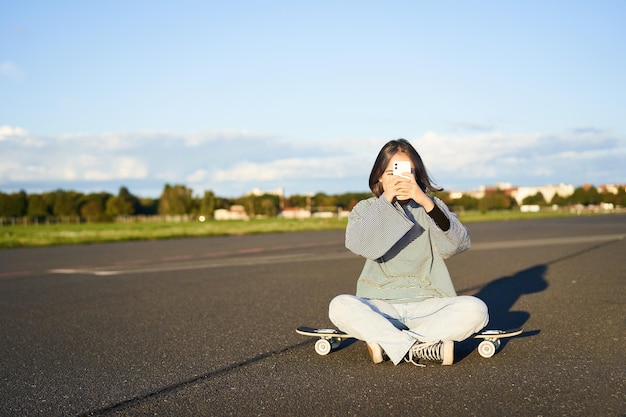 写真 スマートフォンで写真を撮る彼女のスケート ボードに座っている流行に敏感な十代の少女アジアの女性スケーターは l に座っています。