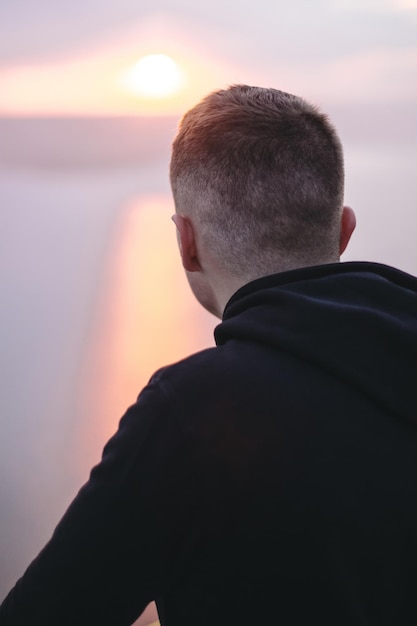 Hipster traveler enjoying amazing sunset view on river while standing on top of rock mountain Brutal guy in windbreaker exploring and traveling Atmospheric moment Copy space