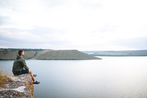 Hipster traveler sitting on top of rock mountain and enjoying amazing view on river Stylish guy exploring and traveling Atmospheric tranquil moment Copy space