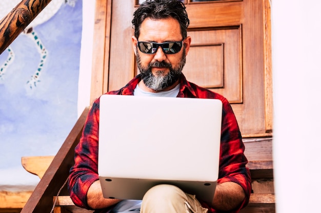 Foto hipster volwassen man met baard gebruik persoonlijke laptop computer buiten thuis zitten op een trap