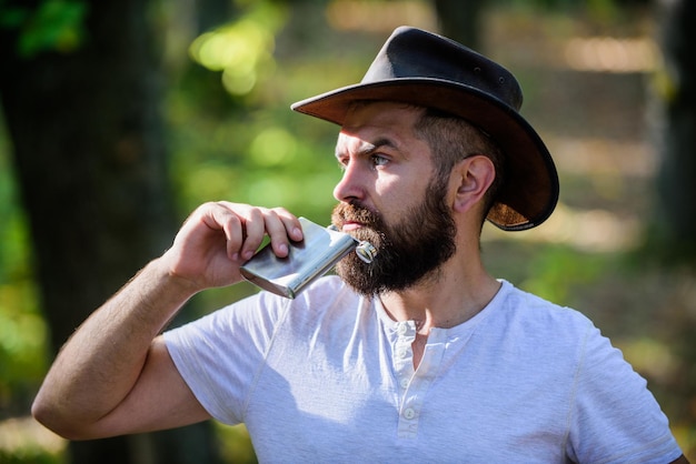 Hipster with beard drink alcohol nature background defocused Hiking camping Wanderer concept Alcohol drink Nomadic man carry alcohol with him Guy brutal bearded cowboy drink alcohol metal flask