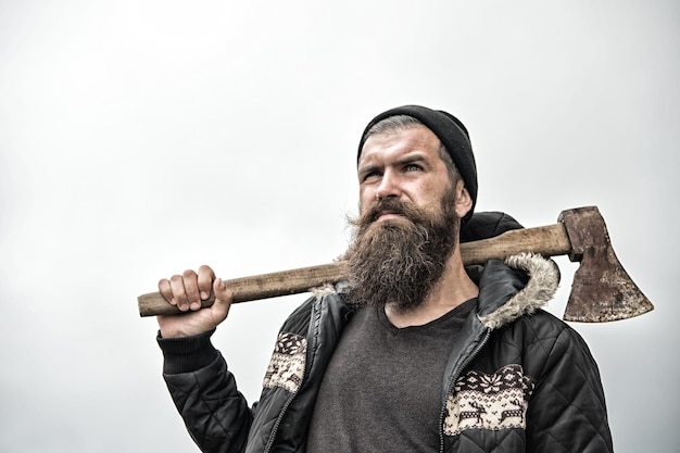 Hipster with beard on serious face carries axe on shoulder sky on background, copy space. Lumberjack brutal and bearded holds axe. Brutal lumberjack concept. Man in hat and warm jacket looks brutally.