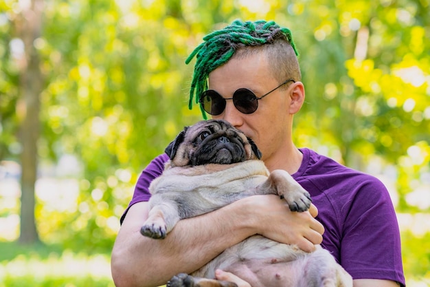 Hipster with green dreadlocks and black round glasses holding and stroking a cute pug on a sunny summer day A man walks his dog